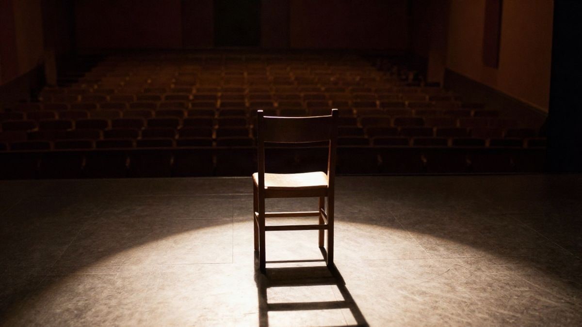 Imagen ilustrativa: una silla de madera en un escenario bajo un foco, con sombras alargadas