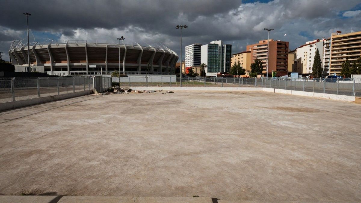 Imagen ilustrativa: una parcela vacía cercada junto a un estadio deportivo y edificios modernos al fondo, con luz natural.