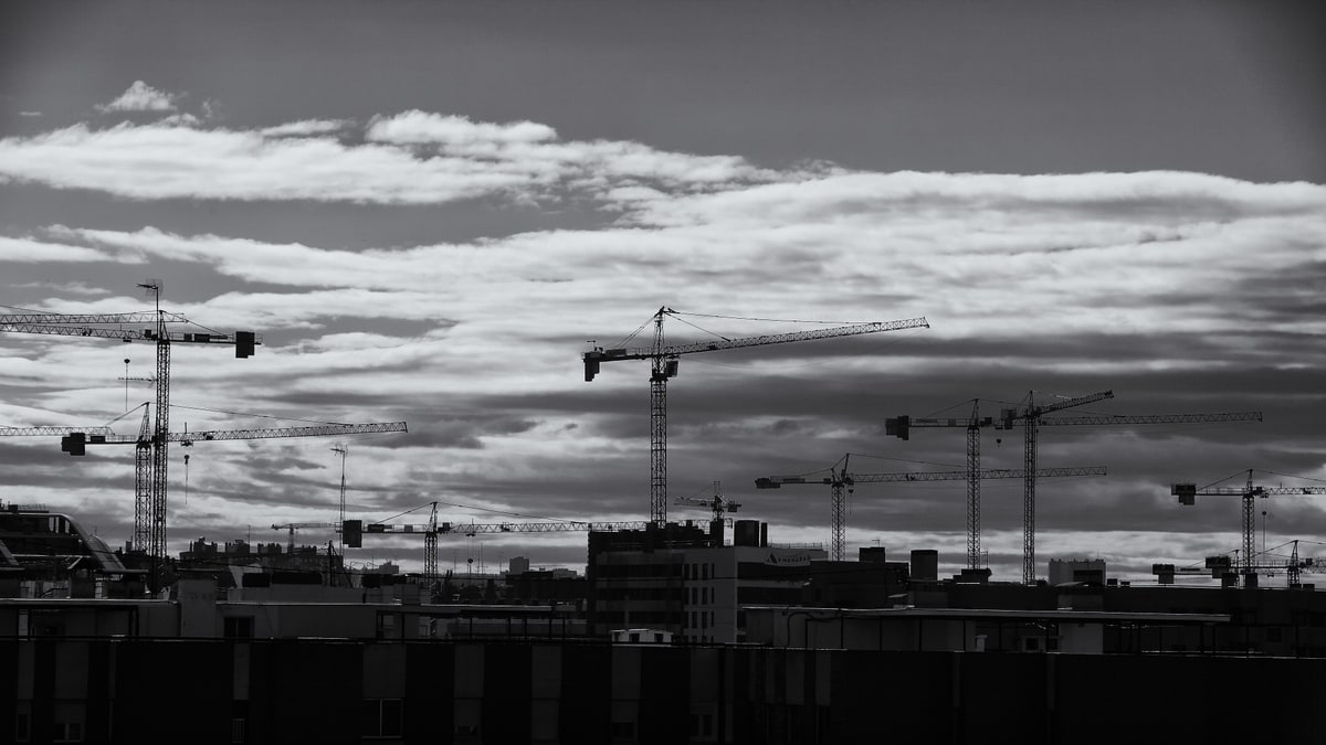 Grúas de construcción recortadas contra un cielo nublado sobre los tejados de una ciudad, en blanco y negro.