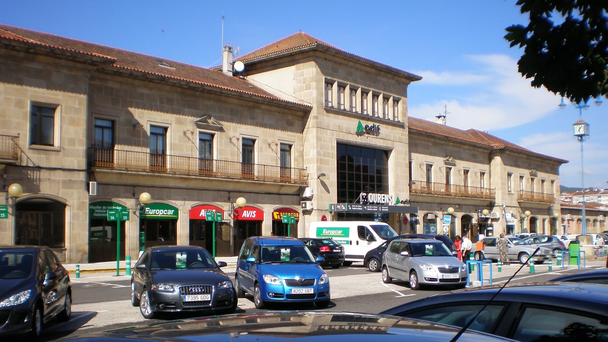 Fachada de la estación de Ourense con coches aparcados y rótulos de empresas de alquiler de vehículos