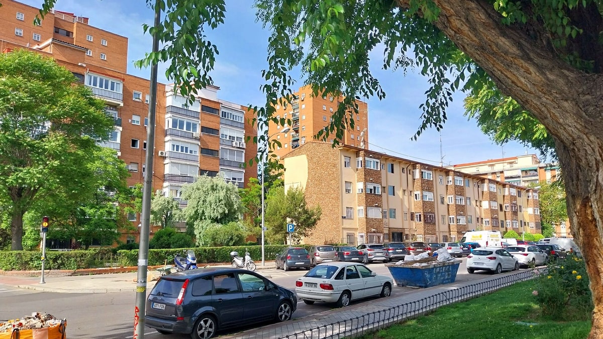 Calle residencial de Carabanchel con bloques de pisos, coches aparcados y contenedores junto a la acera.