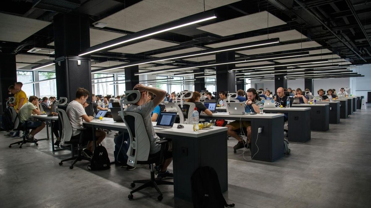 Imagen ilustrativa: aula de informática moderna y vacía, con filas de mesas y monitores que convergen hacia una gran ventana con luz natural