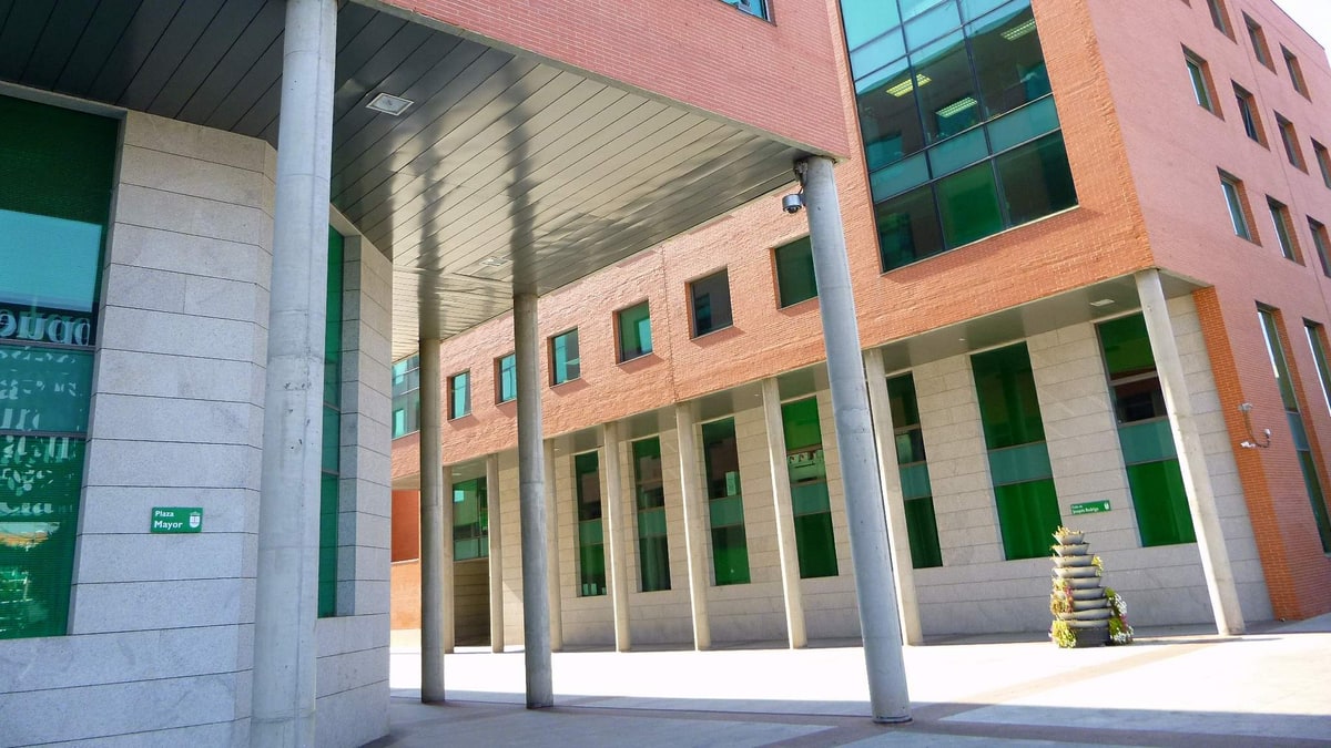 Fachada de ladrillo y cristal con soportales de columnas y cartel verde en la entrada del edificio municipal.