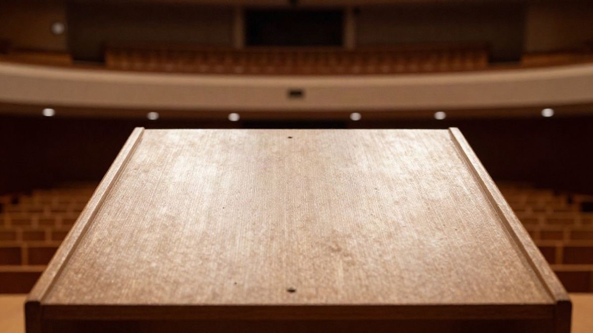 Imagen ilustrativa: primer plano de un atril vacío en un auditorio con butacas desenfocadas al fondo