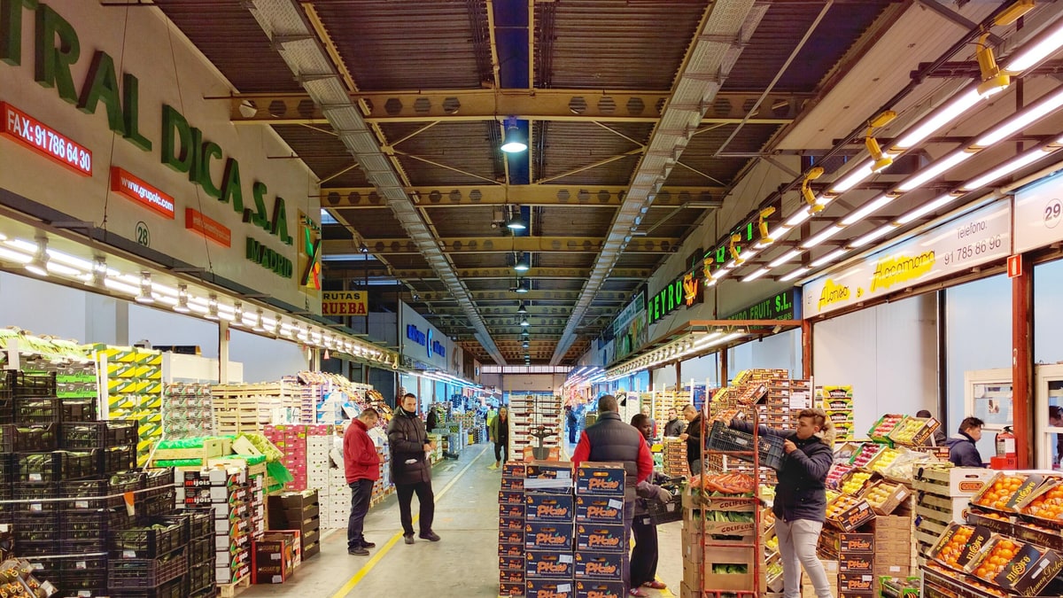 Pasillo del mercado mayorista con cajas de fruta apiladas y compradores entre los puestos