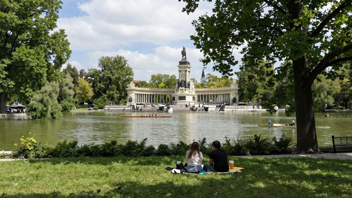 Vista del Parque del Retiro en Madrid, con árboles verdes, un lago y un picnic en la hierba.